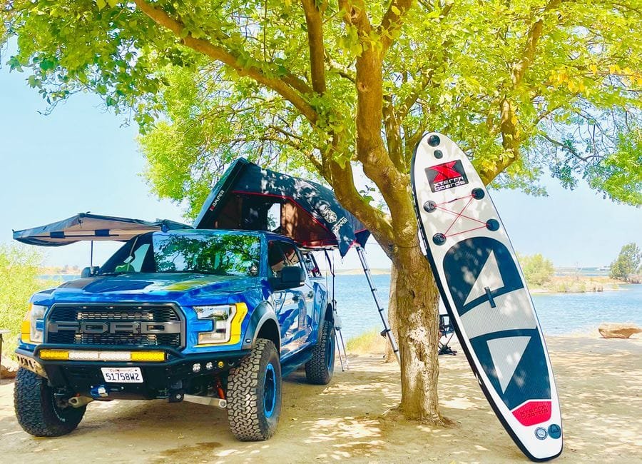 A vibrant scene features an off-road truck, tent, and paddleboard near a scenic lakeside, perfect for outdoor adventure. (Photo by Tiffany Bacon on Pexels)