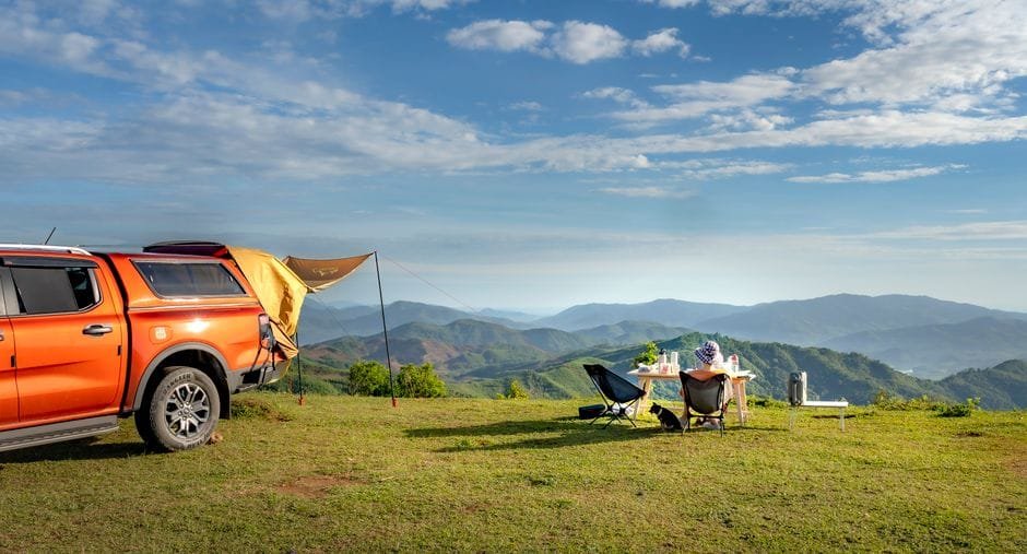 Outdoor camping setup with chairs and table beside orange pick-up truck on hilltop, overlooking mountains. (Photo by Quang Nguyen Vinh on Pexels)