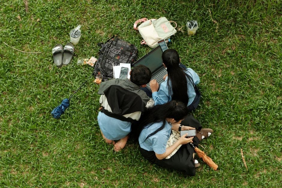 Top view of three friends using a laptop and phones on grass, enjoying a picnic outdoors. (Photo by Sophanith CHHOUR on Pexels)