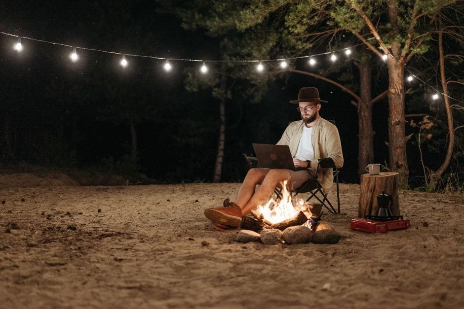 Digital nomad working remotely by a campfire with a laptop under string lights in the forest. (Photo by Pavel Danilyuk on Pexels)