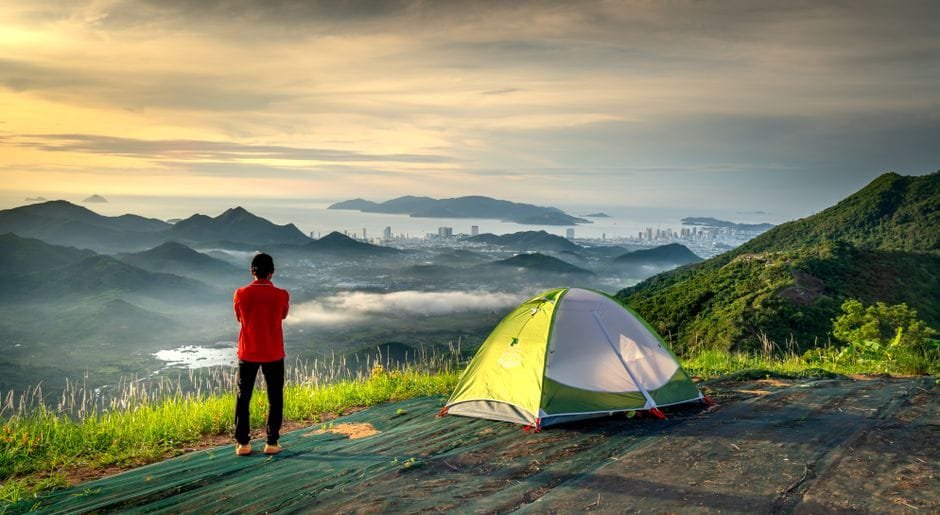 Adventurer stands by tent, embracing scenic mountain and city view at sunrise. (Photo by Quang Nguyen Vinh on Pexels)