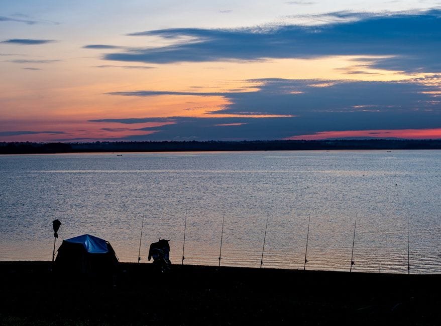 Tranquil lakeside view at sunset with a tent and fishing rods on the shore. (Photo by rick mun on Pexels)