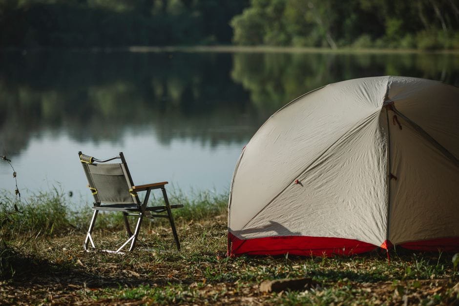 Tent and folding chair on grass coast against trees reflecting in lake in soft sunlight (Photo by Uriel Mont on Pexels)