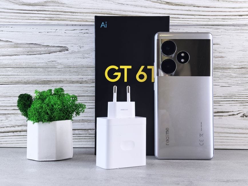 Close-up of a Realme smartphone with charger and potted plant on a wooden surface. (Photo by Andrey Matveev on Pexels)