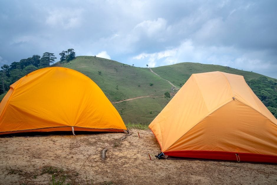 Colorful orange camping tents located on sandy ground near green mountain covered with grass against cloudy sky on summer day (Photo by Quang Nguyen Vinh on Pexels)