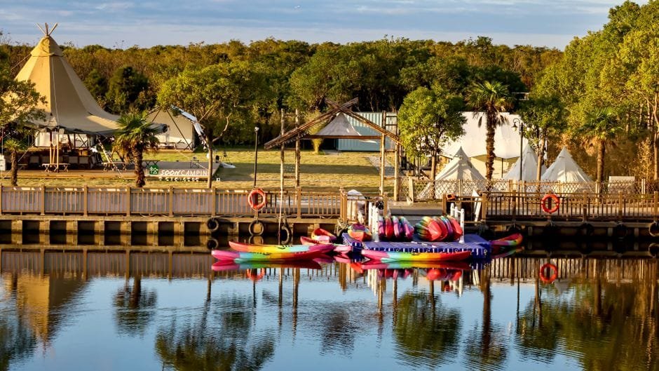 Colorful kayaks docked by a scenic campground by the lake with tents and lush greenery. (Photo by Yudi Ding on Pexels)