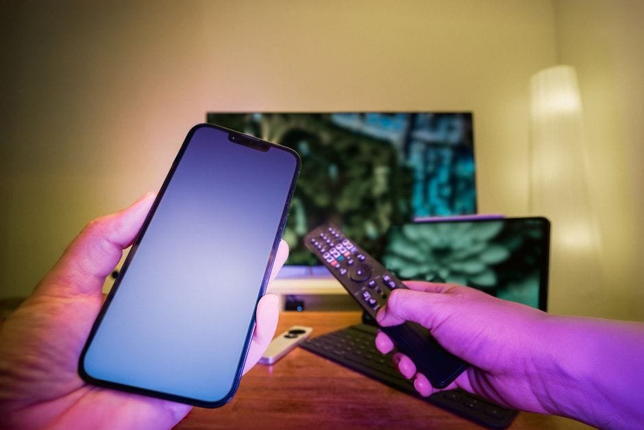 Hands holding a smartphone and remote control in a high-tech home setup with TV and other electronic devices. (Photo by Jakub Zerdzicki on Pexels)