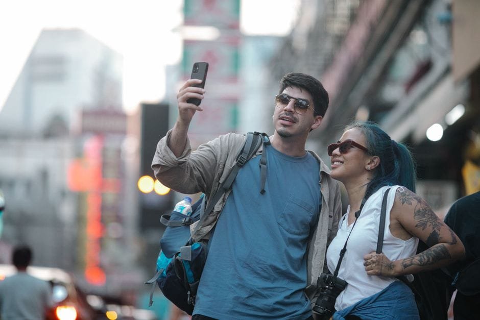 Smiling couple capturing a selfie on a busy city street, enjoying travel moments. (Photo by George Pak on Pexels)