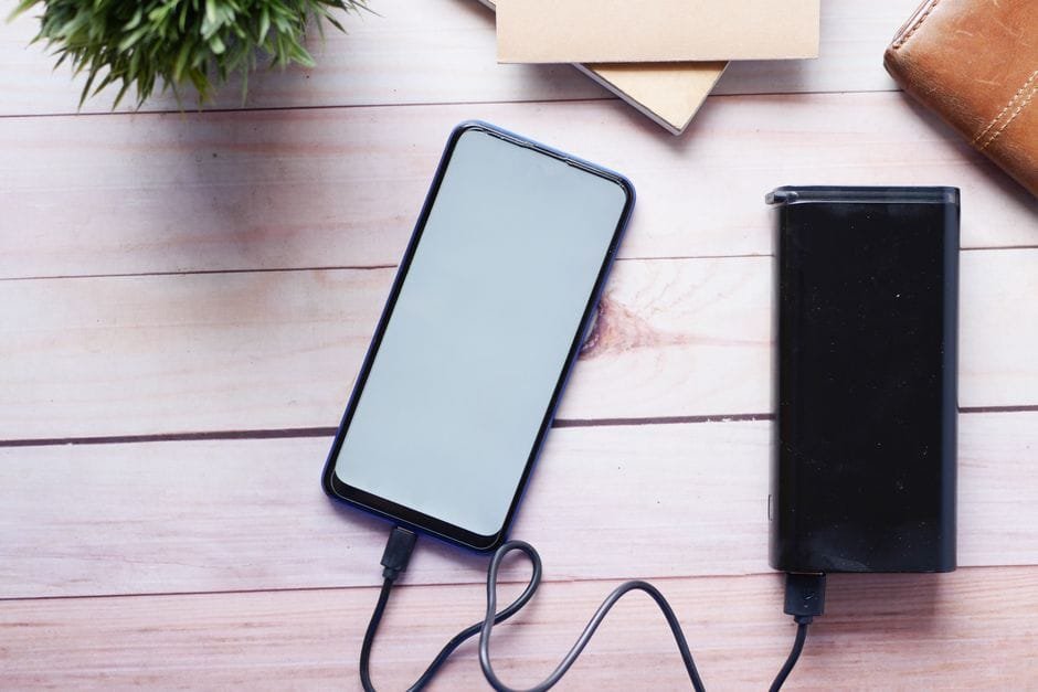 Smartphone charging with a power bank on a wooden desk, showcasing modern technology essentials. (Photo by Towfiqu barbhuiya on Pexels)
