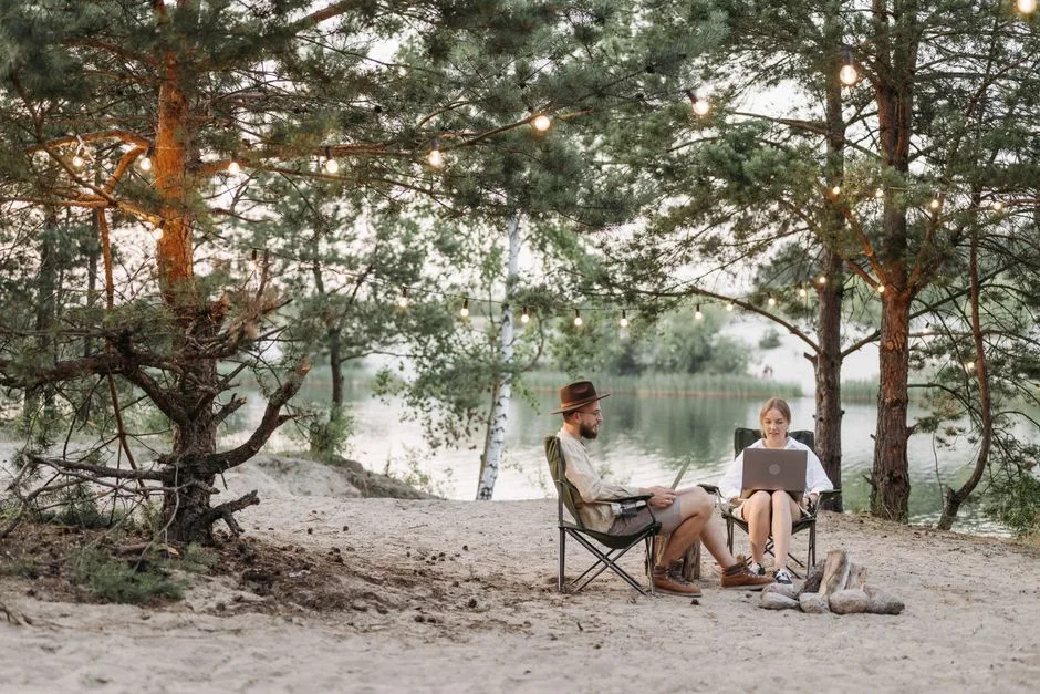 A couple enjoys a serene camping experience by a lake, working on a laptop under string lights. (Photo by Pavel Danilyuk on Pexels)