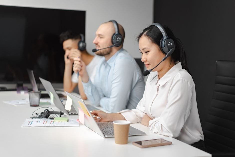Call center agents wearing headsets working on laptops at a customer support team meeting. (Photo by Mikhail Nilov on Pexels)