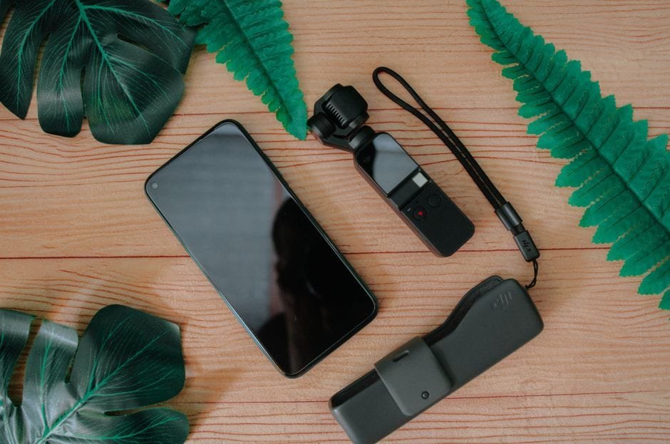 Top view of mobile phone and mini handheld camera with flash drive composed on wooden table with green leaves of tropical plants (Photo by Pok Rie on Pexels)