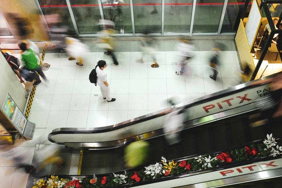 Blurred motion of people moving on escalator in busy transit hub with floral decorations. (Photo by Mace Pacardo on Pexels)