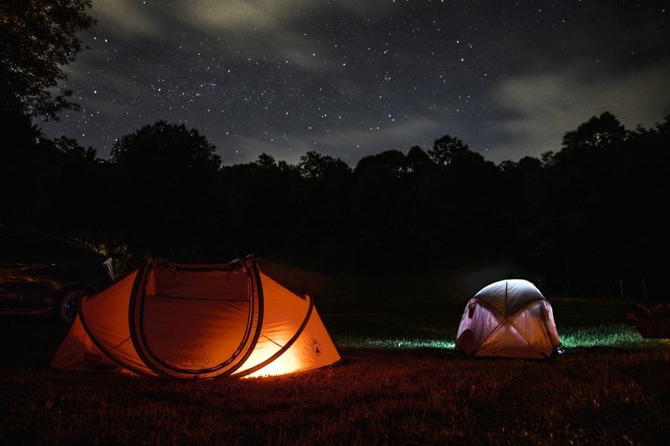 A serene night camping scene with glowing tents under a starlit sky. (Photo by Matthew DeVries on Pexels)