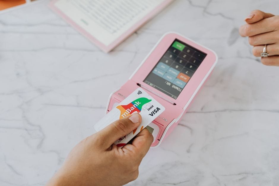 Hand using a contactless card on a pink POS terminal for a wireless transaction. (Photo by https://kaboompics.com/ on Pexels)