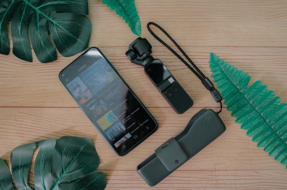 A flat lay of a smartphone and camera with green leaves on a wooden table. (Photo by Pok Rie on Pexels)