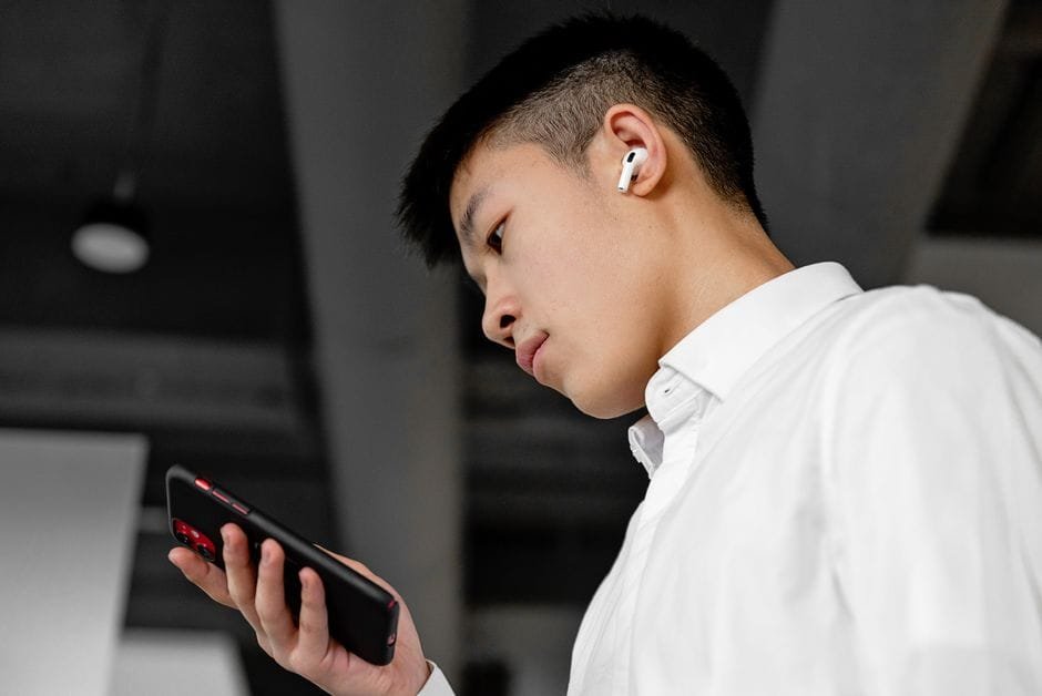 Side view of a young man wearing wireless earbuds and using a smartphone indoors. (Photo by Mikhail Nilov on Pexels)